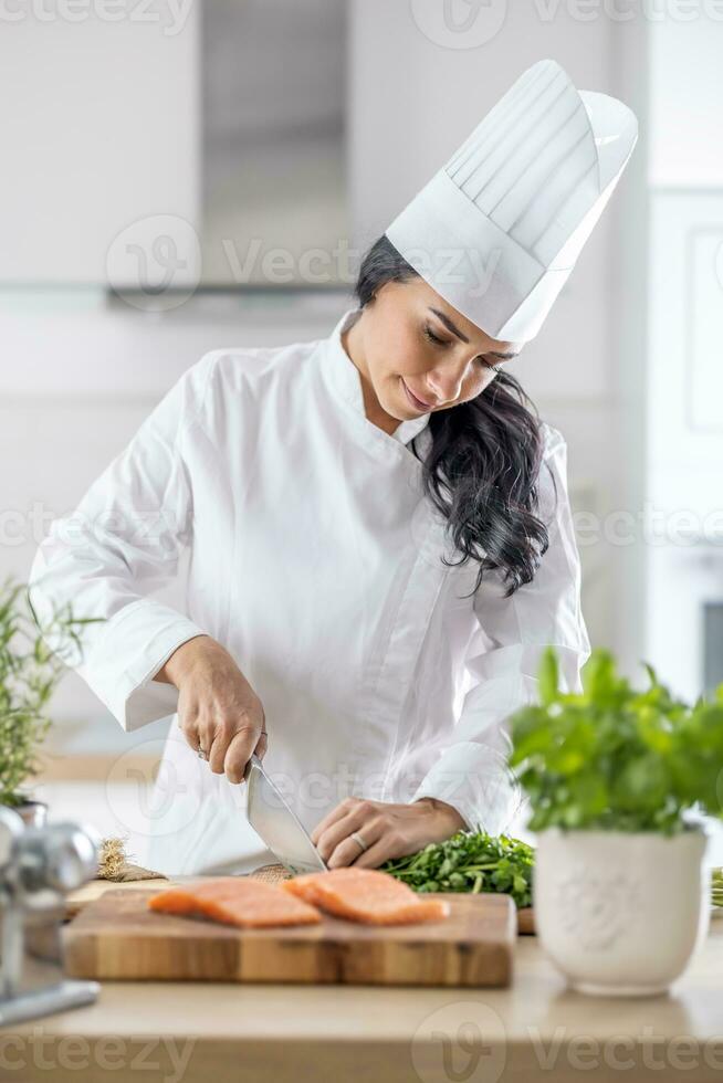 felice, messa a fuoco, professionale femmina capocuoco corre coltello attraverso fresco filetto di pesce con fresco erbe aromatiche su il lato foto