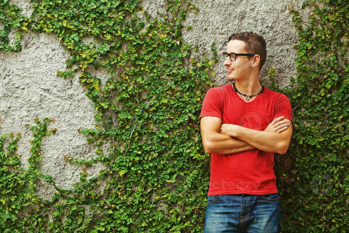 un' uomo nel un' rosso camicia e bicchieri in piedi nel davanti di un' parete coperto nel edera foto