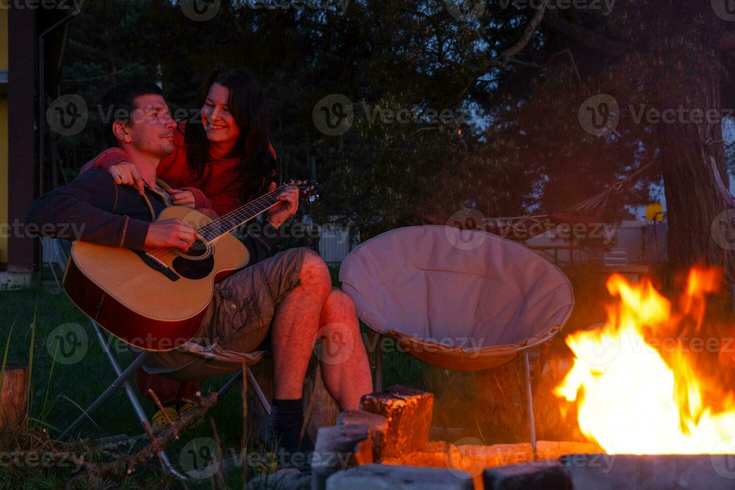 un' uomo giochi il chitarra, un' donna ascolta e canta lungo. un' coppia nel amore è seduta di il all'aperto fuoco di bivacco nel il cortile di il Casa su campeggio sedie, un' romantico sera foto