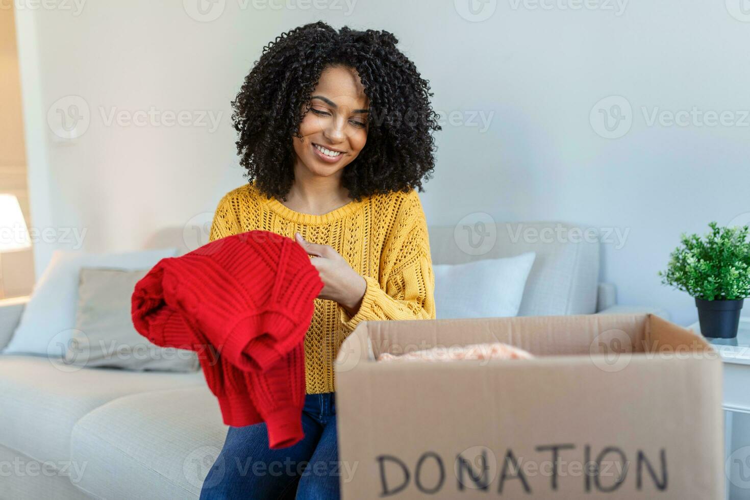 contento africano giovane donna sedersi su divano incollato Abiti nel donazione scatola a casa, cura birazziale femmina volontario mettere abbigliamento nel scatola di cartone pacchetto, donare per bisognoso le persone, riutilizzare, riciclare concetto foto