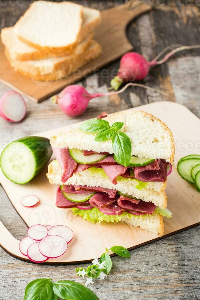 Doppio Sandwich con pastrami e fresco verdure e erbe aromatiche su un' taglio tavola. americano merenda. rustico stile. verticale Visualizza foto