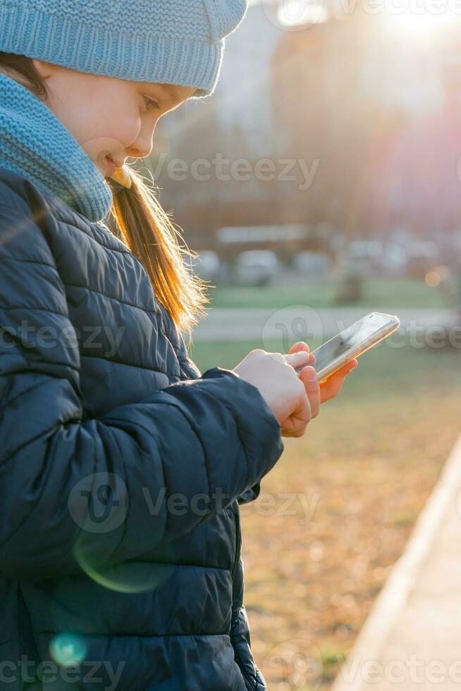 un' ragazza nel un' giacca è Tenere un' smartphone e sms un' Messaggio nel il raggi di il sera sole. Internet fare surf foto