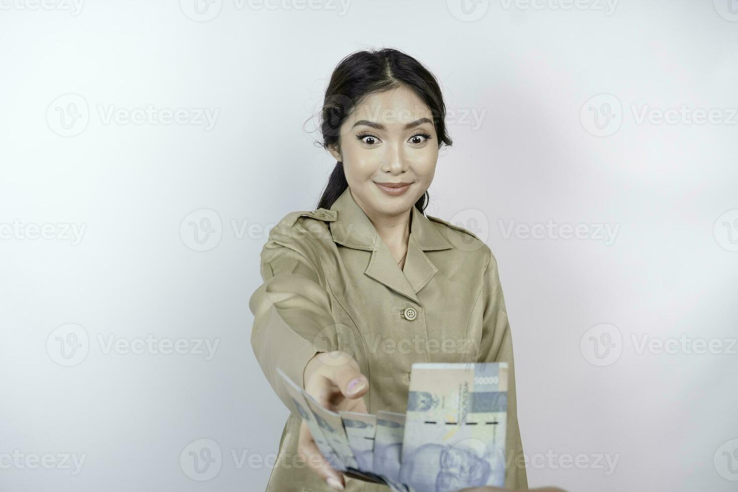 un' sorridente giovane governo lavoratore donna è indossare cachi uniforme Tenere denaro contante i soldi nel indonesiano rupia isolato di bianca sfondo foto