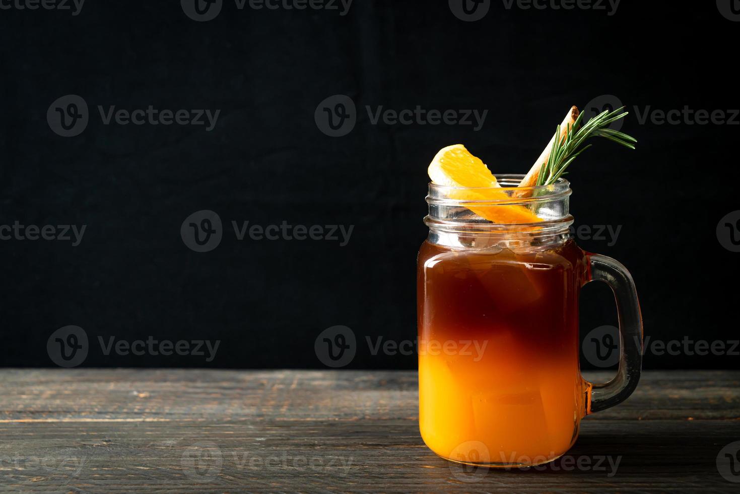 un bicchiere di caffè nero americano ghiacciato e uno strato di succo di arancia e limone decorato con rosmarino e cannella su uno sfondo di legno foto