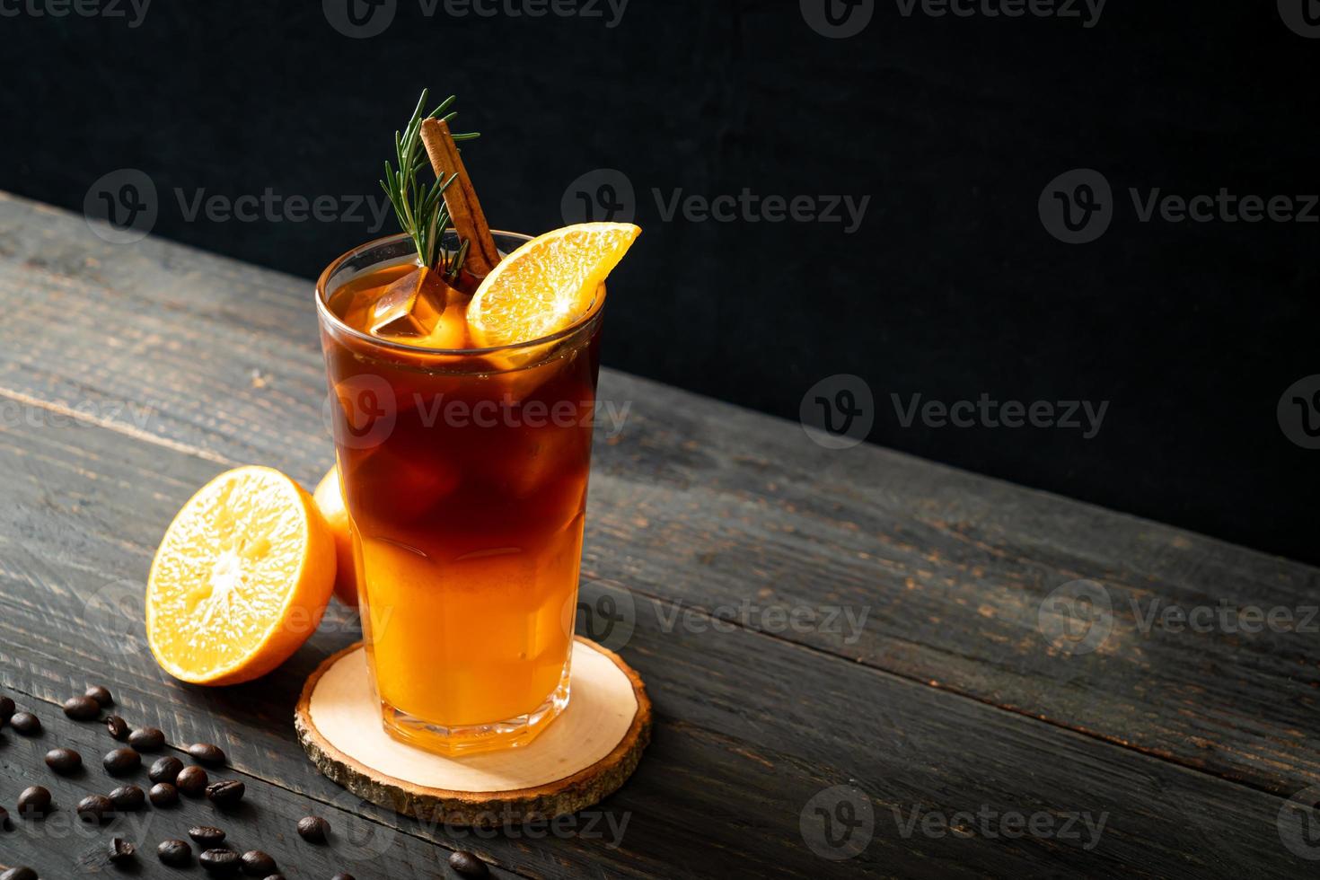 un bicchiere di caffè nero americano ghiacciato e uno strato di succo di arancia e limone decorato con rosmarino e cannella su uno sfondo di legno foto