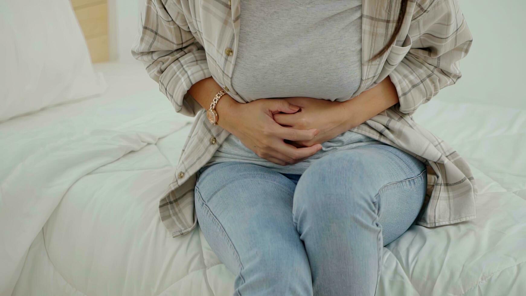 asiatico giovane donna sofferenza a partire dal addominale dolore mentre seduta nel letto a casa. foto