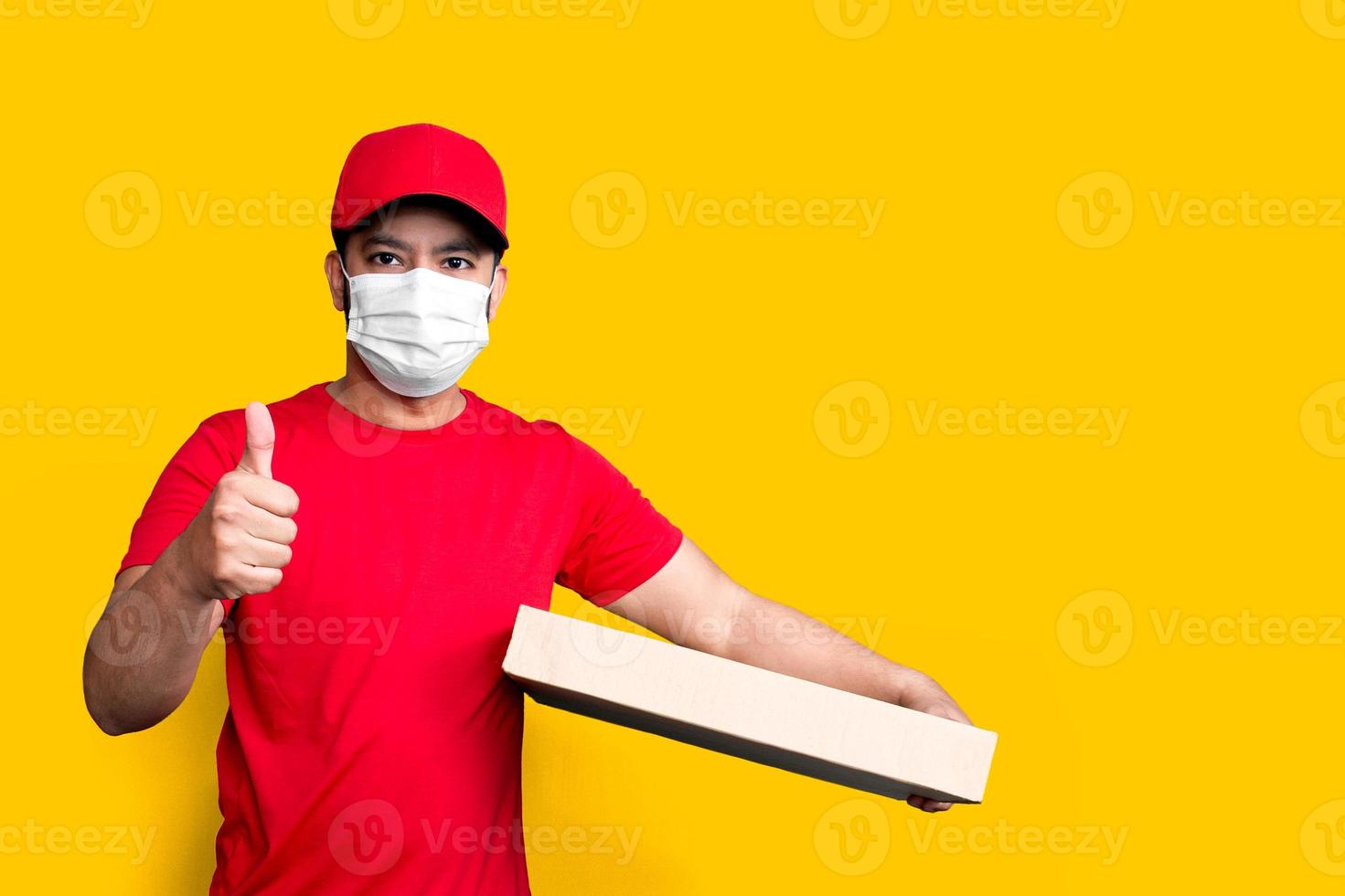 impiegato dell'addetto alle consegne con berretto rosso maglietta bianca uniforme maschera per il viso tenere una scatola di cartone vuota isolata su sfondo giallo foto