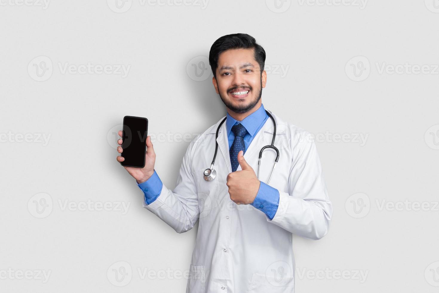 medico che tiene il telefono che mostra pollice in su isolato. uomo pakistano medico tecnologia medicina a casa. schermo chiaro del telefono. foto