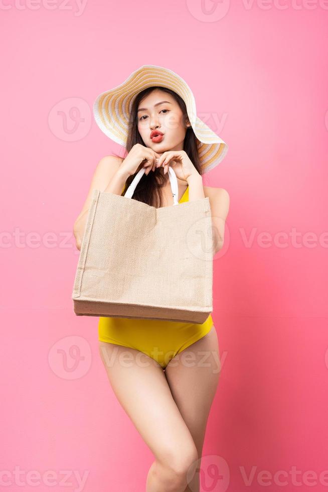 bella giovane donna asiatica in costume da bagno giallo, borsa e cappello a tesa larga in posa su sfondo rosa foto