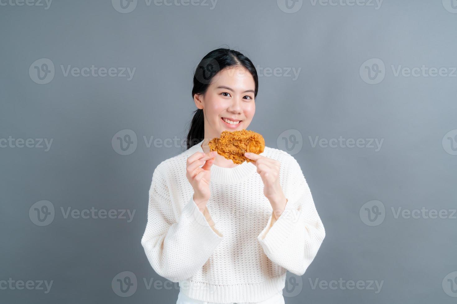 giovane donna asiatica che indossa un maglione con una faccia felice e si diverte a mangiare pollo fritto su sfondo grigio foto