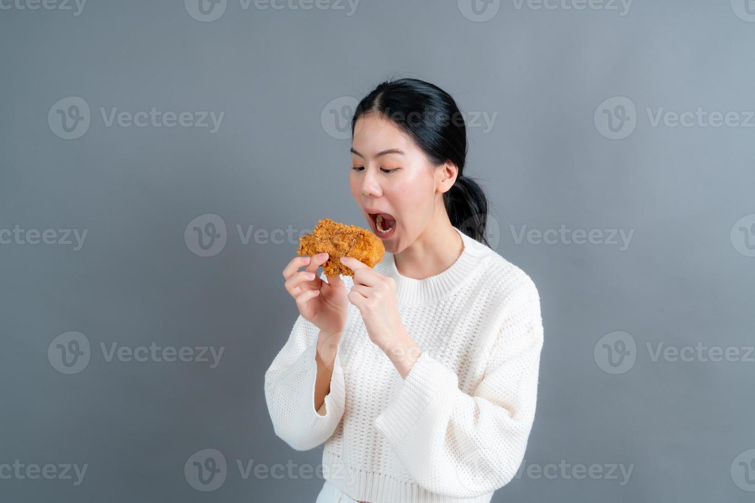 giovane donna asiatica che indossa un maglione con una faccia felice e si diverte a mangiare pollo fritto su sfondo grigio foto