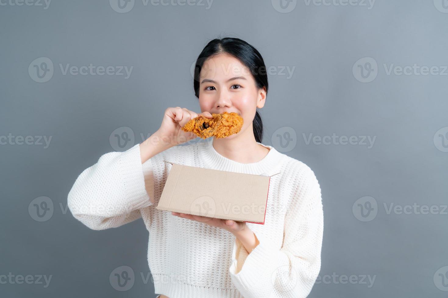 giovane donna asiatica che indossa un maglione con una faccia felice e si diverte a mangiare pollo fritto su sfondo grigio foto