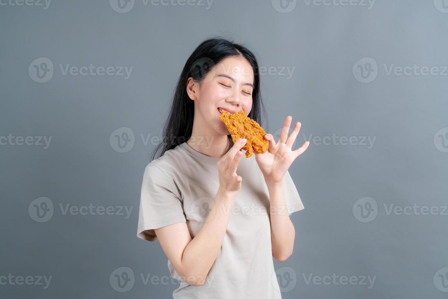 giovane donna asiatica che indossa un maglione con una faccia felice e si diverte a mangiare pollo fritto su sfondo grigio foto