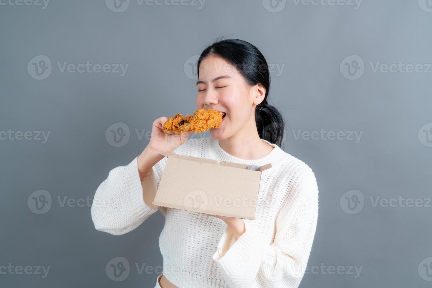 giovane donna asiatica che indossa un maglione con una faccia felice e si diverte a mangiare pollo fritto su sfondo grigio foto