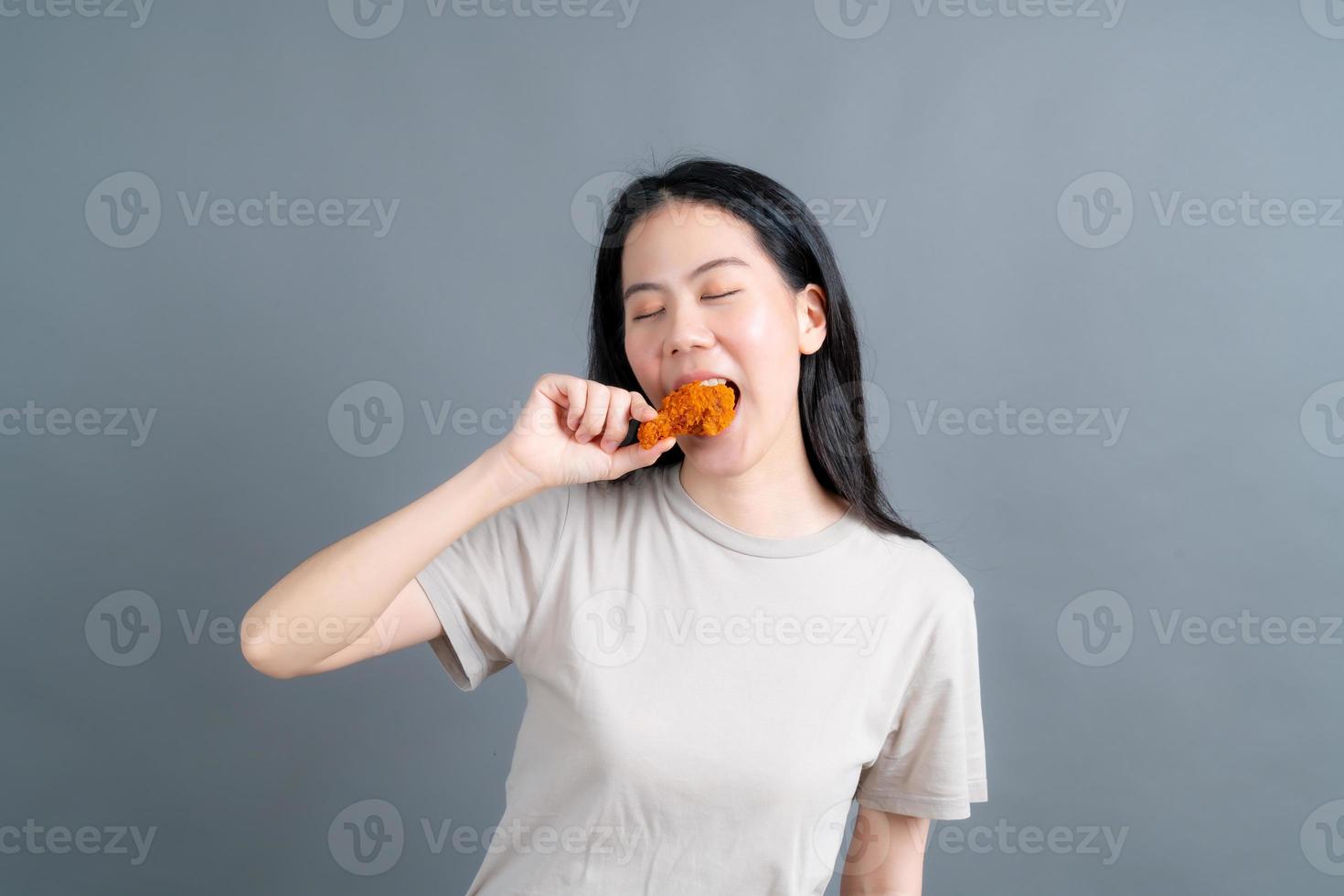 giovane donna asiatica che indossa un maglione con una faccia felice e si diverte a mangiare pollo fritto su sfondo grigio foto