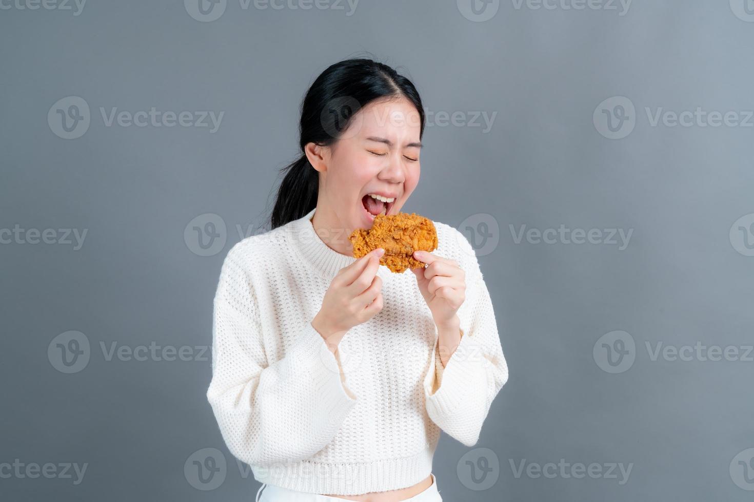 giovane donna asiatica che indossa un maglione con una faccia felice e si diverte a mangiare pollo fritto su sfondo grigio foto