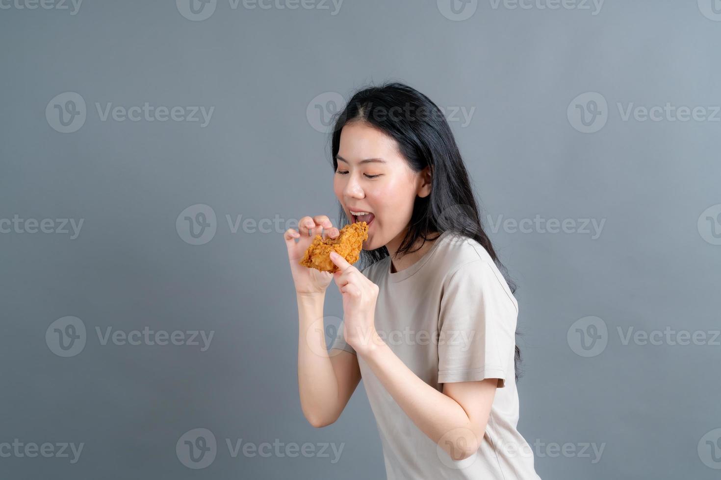 giovane donna asiatica che indossa un maglione con una faccia felice e si diverte a mangiare pollo fritto su sfondo grigio foto