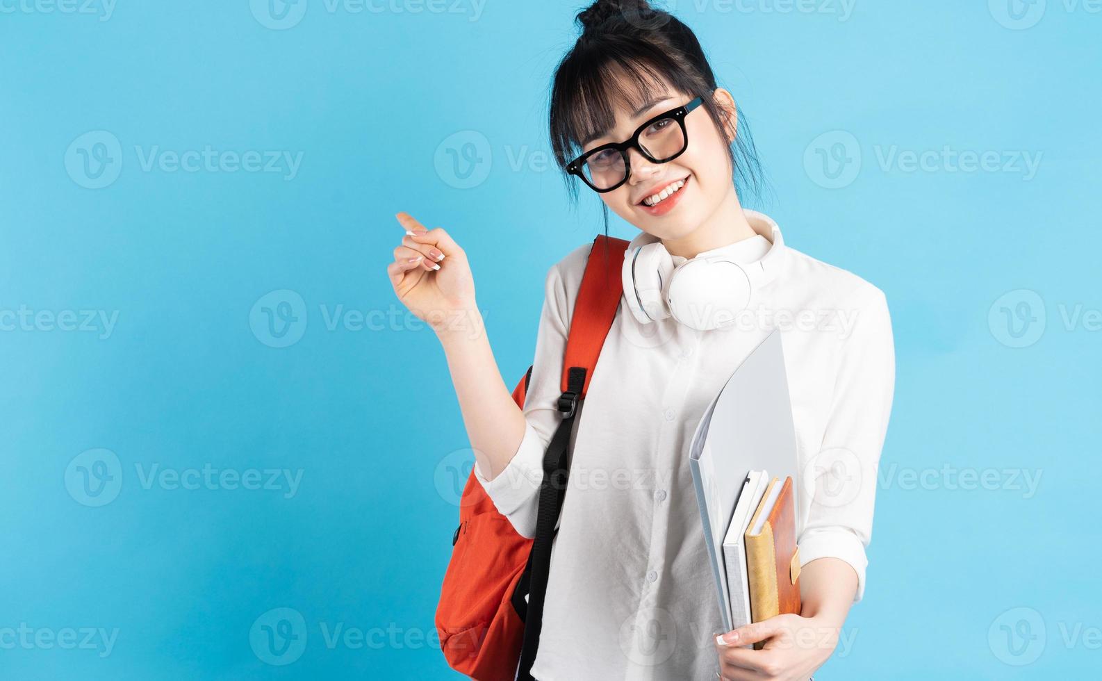 studentessa asiatica che indossa uno zaino dietro la schiena, tiene in mano uno smartphone, indossa al collo auricolari wireless, tiene in mano un bicchiere di carta foto