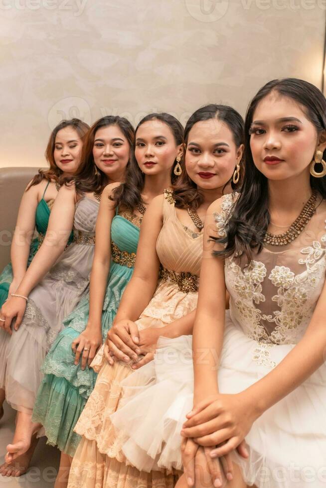 un' gruppo di asiatico donne seduta insieme su un' bianca letto mentre indossare vestiti e trucco durante un' festa a un' gli amici Casa foto