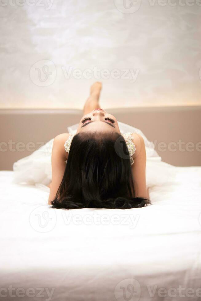 un asiatico donna nel un' bianca vestito è dire bugie su un' bianca letto con sua nero capelli perdere nel un' Hotel foto
