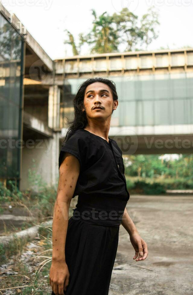 un asiatico uomo con lungo capelli e indossare nero Abiti in piedi in giro un abbandonato edificio foto