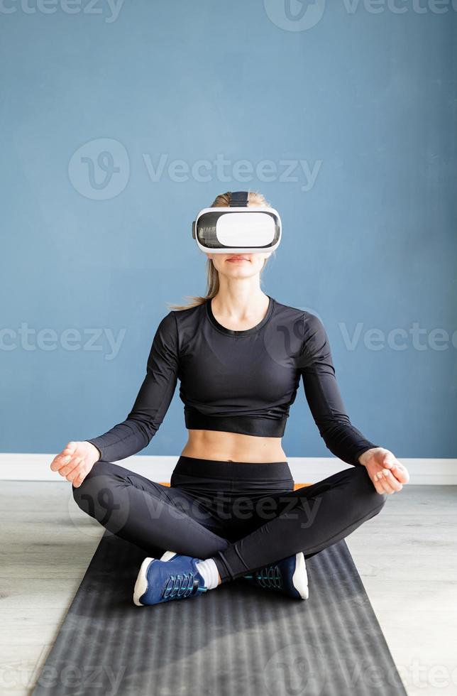 giovane donna bionda in abiti sportivi che indossano occhiali per realtà virtuale meditando sul tappetino fitness foto