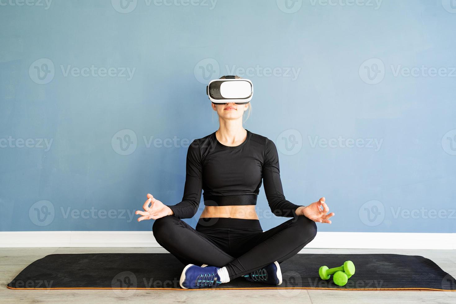 giovane donna bionda in abiti sportivi che indossano occhiali per realtà virtuale meditando sul tappetino fitness foto