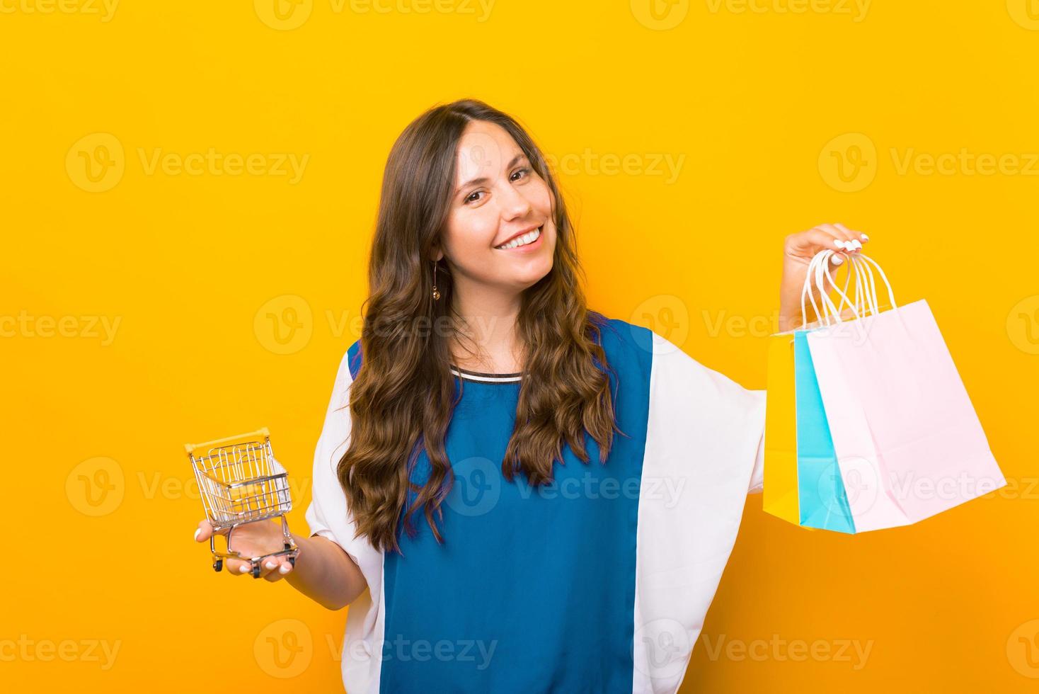 affascinante giovane donna in piedi su sfondo giallo e tenendo borse della spesa e carrello della spesa foto