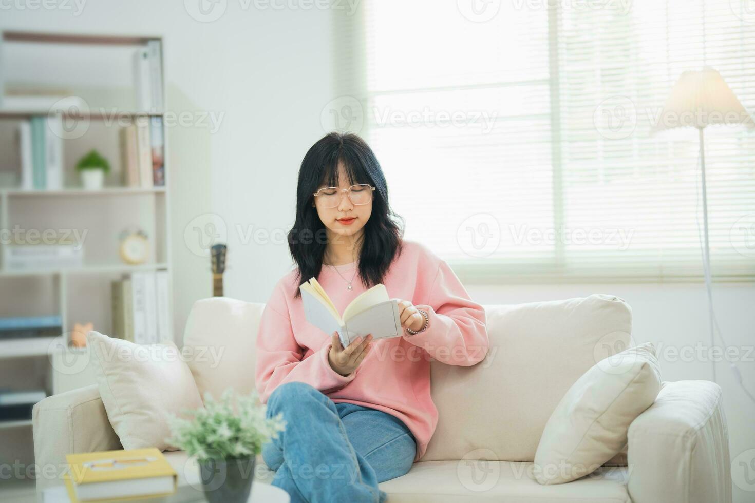 gioioso contento asiatico donna sorridente e lettura libro mentre seduta su divano divano nel vivente camera a casa. asiatico donne rilassare lettura libro Sorridi a divano nel il Casa. attività passatempo a Casa concetto. foto