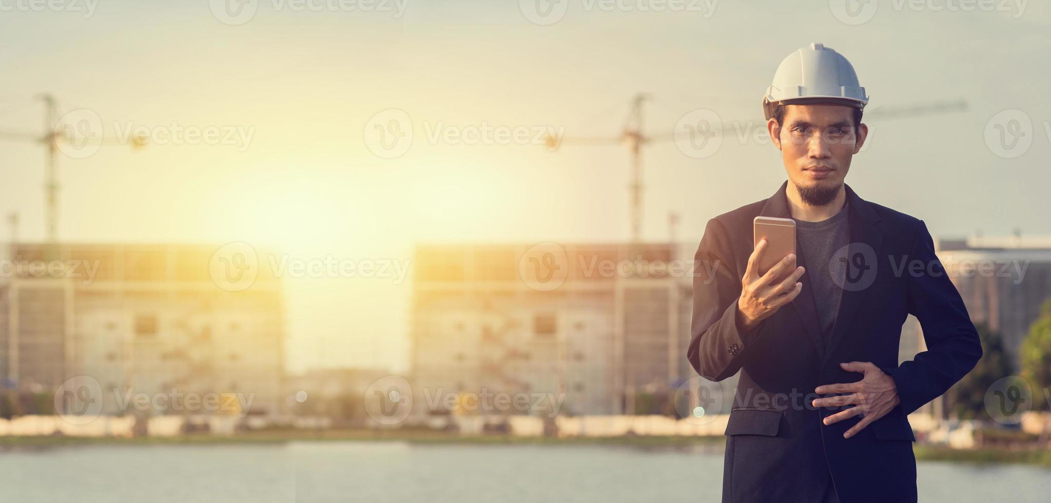 l'uomo d'affari indossa la sicurezza del casco utilizzando il telefono cellulare all'aperto sfondo del sito in costruzione foto