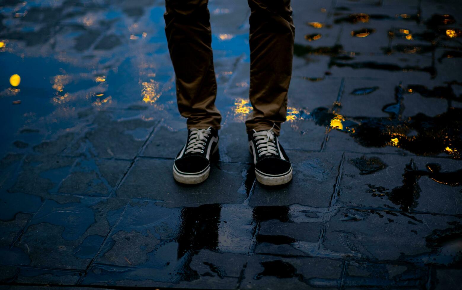 in piedi magro giovane dell'uomo gambe riflessa nel bulboso, circolare bokeh di acqua piovana intrappolati su il cemento pavimento. nel il solitario notte foto