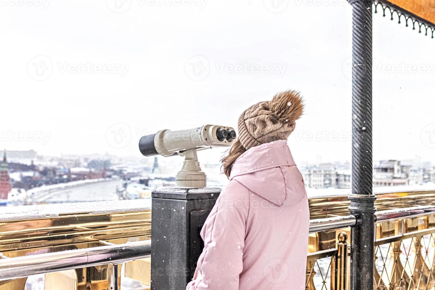 una giovane ragazza guarda attraverso un binocolo a gettoni sul ponte di osservazione che domina la città dall'alto al tramonto. inverno, nevicata foto