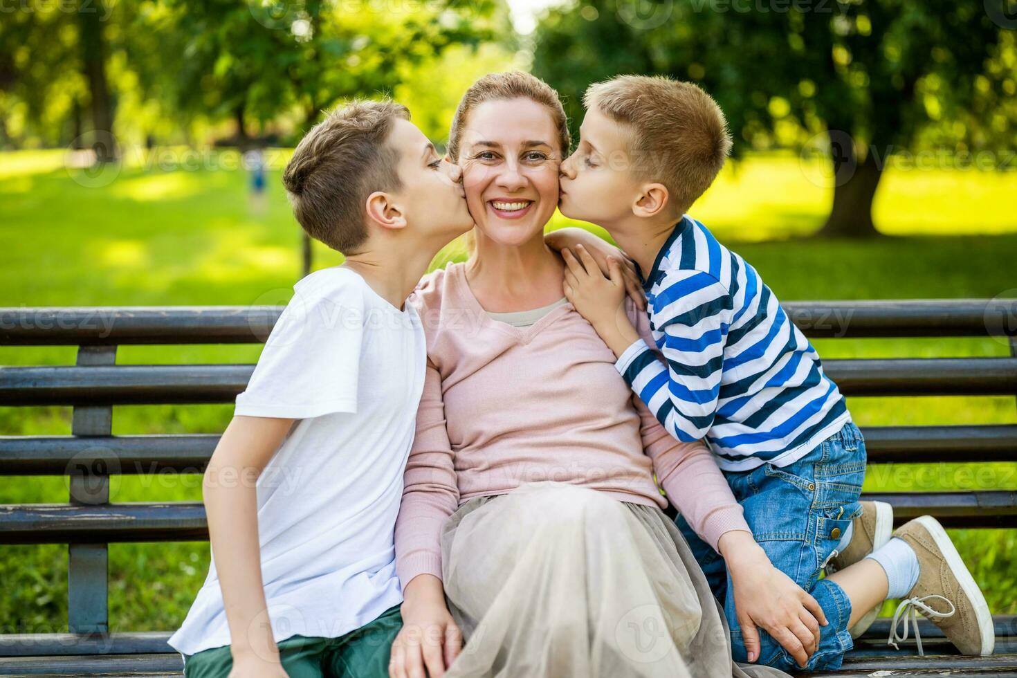 contento madre è seduta con sua figli maschi su panchina nel parco. essi siamo avendo divertimento insieme. ragazzi siamo baci loro madre. foto