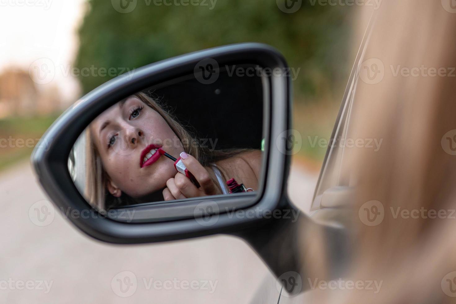 una giovane e bella donna con i capelli lunghi guarda nello specchietto retrovisore dell'auto e si dipinge le labbra. foto