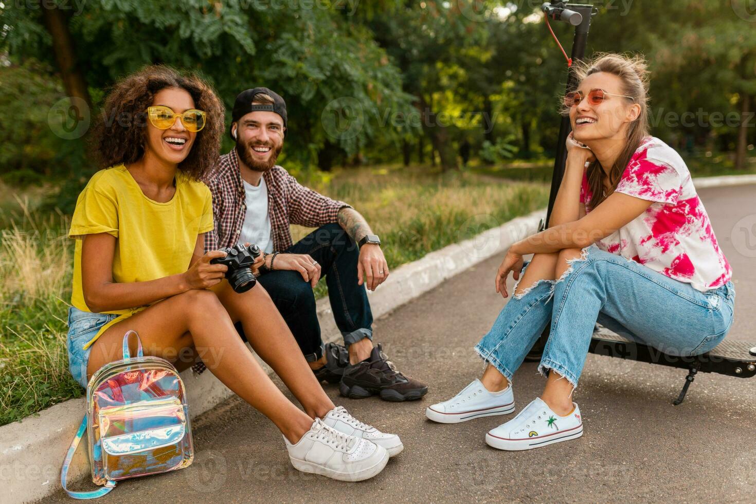 contento giovane azienda di sorridente amici seduta nel parco su erba con elettrico calcio scooter foto