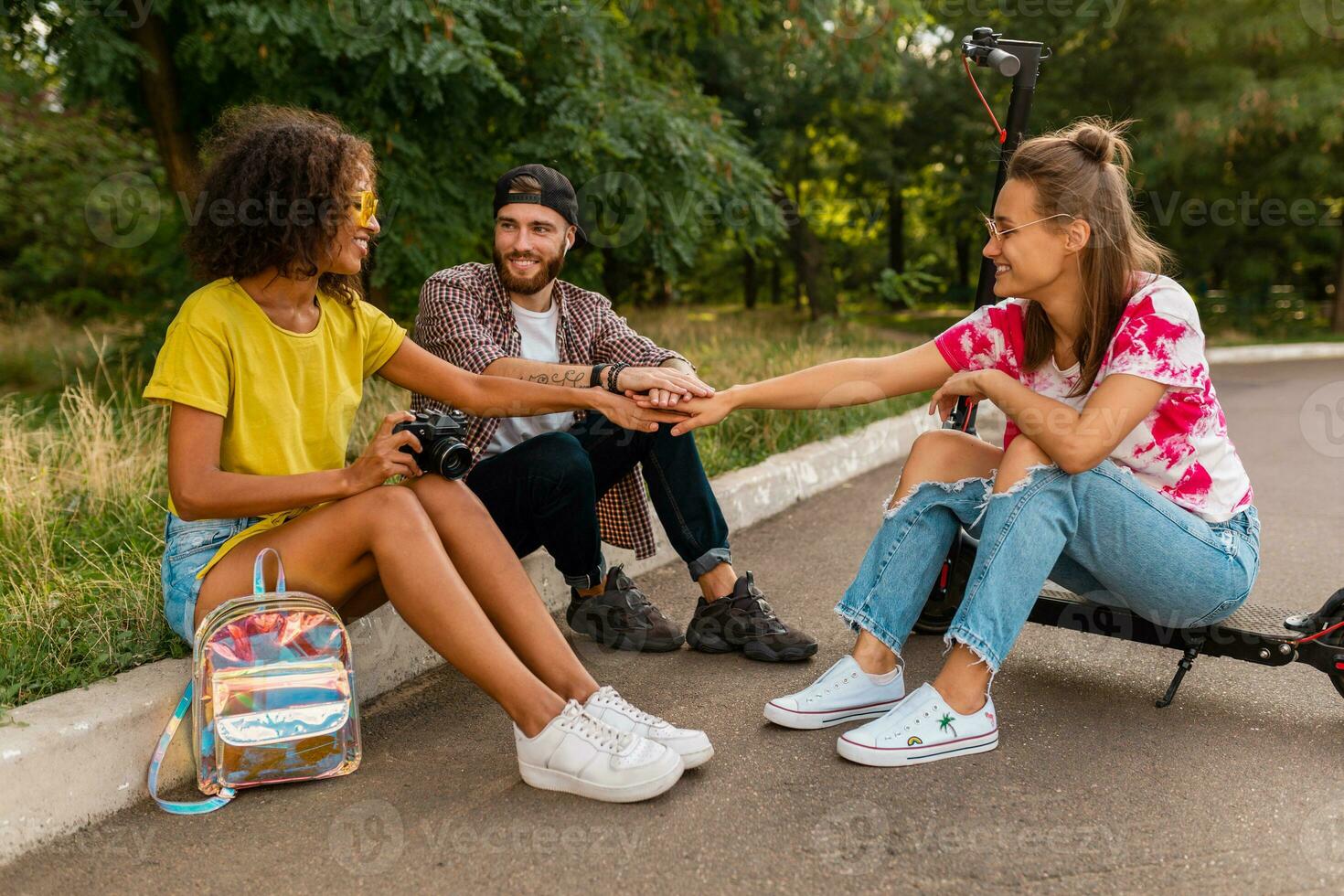 contento giovane azienda di sorridente amici seduta nel parco su erba con elettrico calcio scooter foto