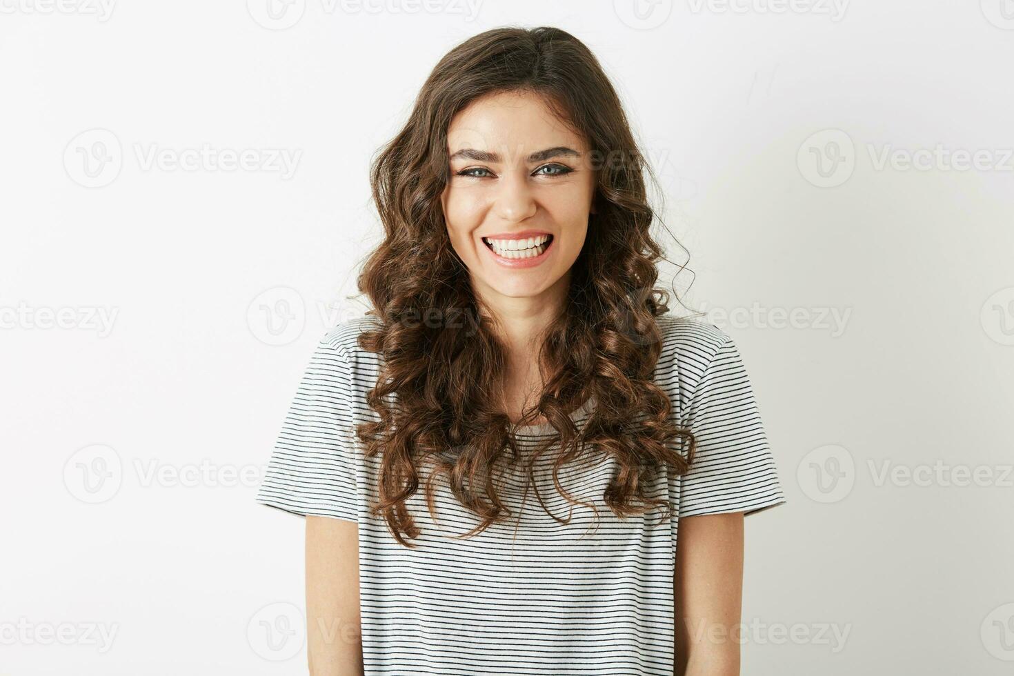 vicino su ritratto di Candido sorridente attraente donna con bianca denti guardare nel telecamera isolato su bianca sfondo, lungo Riccio capelli, semplice maglietta, casuale fricchettone stile, contento positivo emozione foto