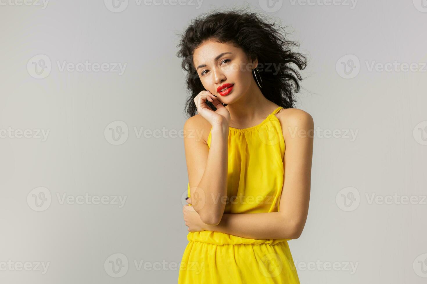 ritratto di giovane sexy brunetta donna nel giallo vestire, toccante sua viso, rosso labbra, Riccio capelli agitando nel vento, estate stile, moda tendenza, guardare nel telecamera, misto gara, isolato, Tenere mani su foto