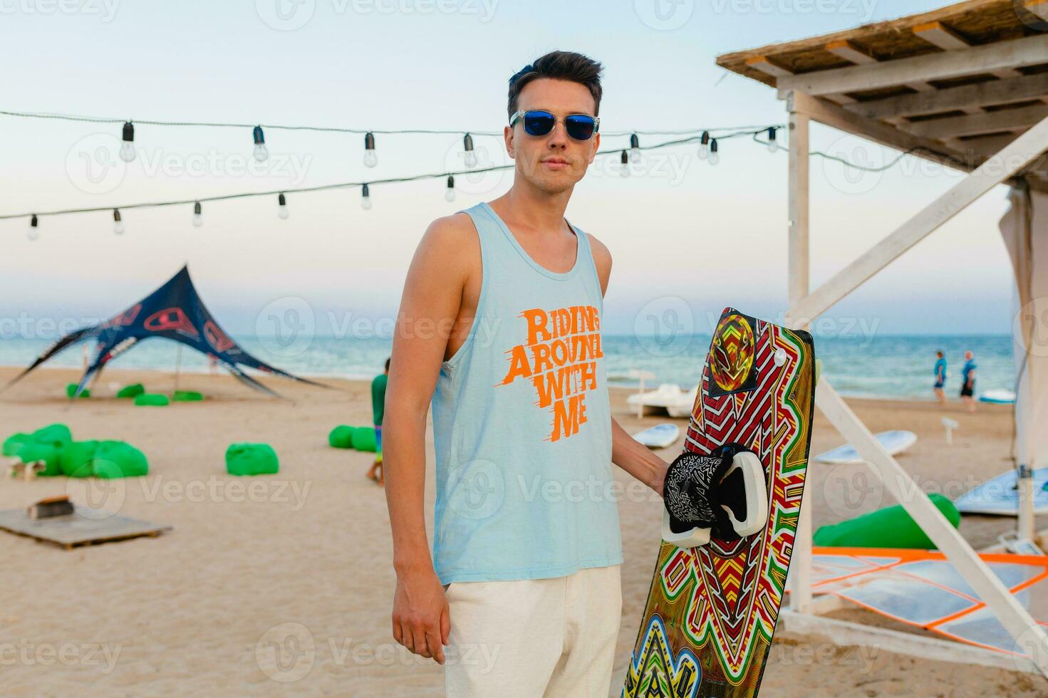 giovane atletico uomo con aquilone fare surf tavola in posa su spiaggia indossare occhiali da sole su estate vacanza, attivo gli sport passatempo foto