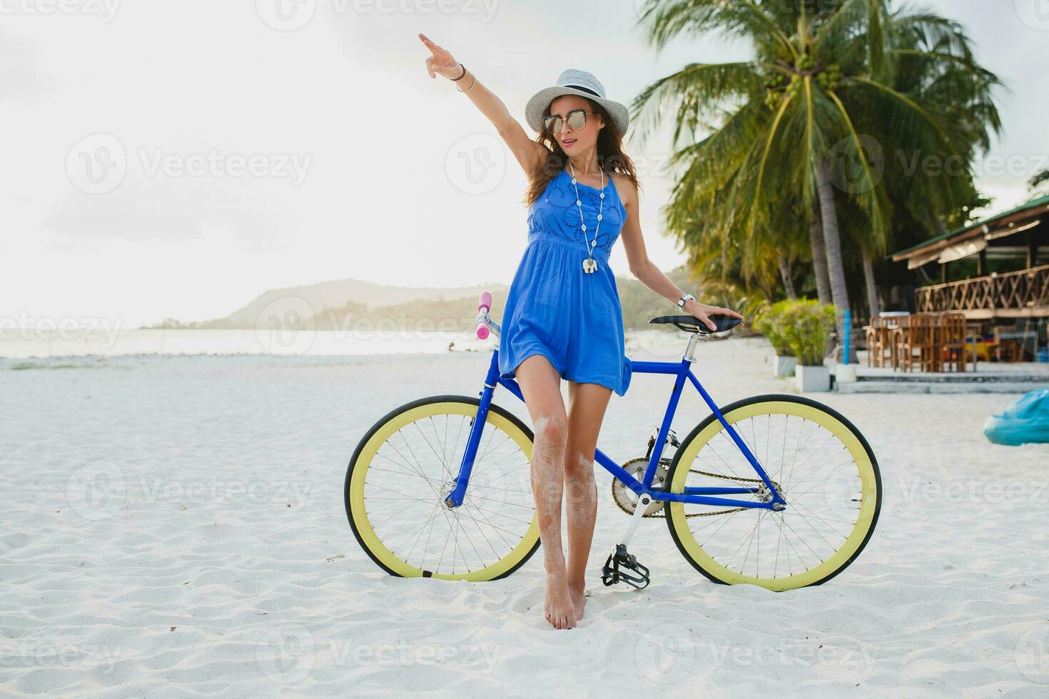 giovane attraente sorridente donna nel blu vestito a piedi su tropicale spiaggia con bicicletta foto