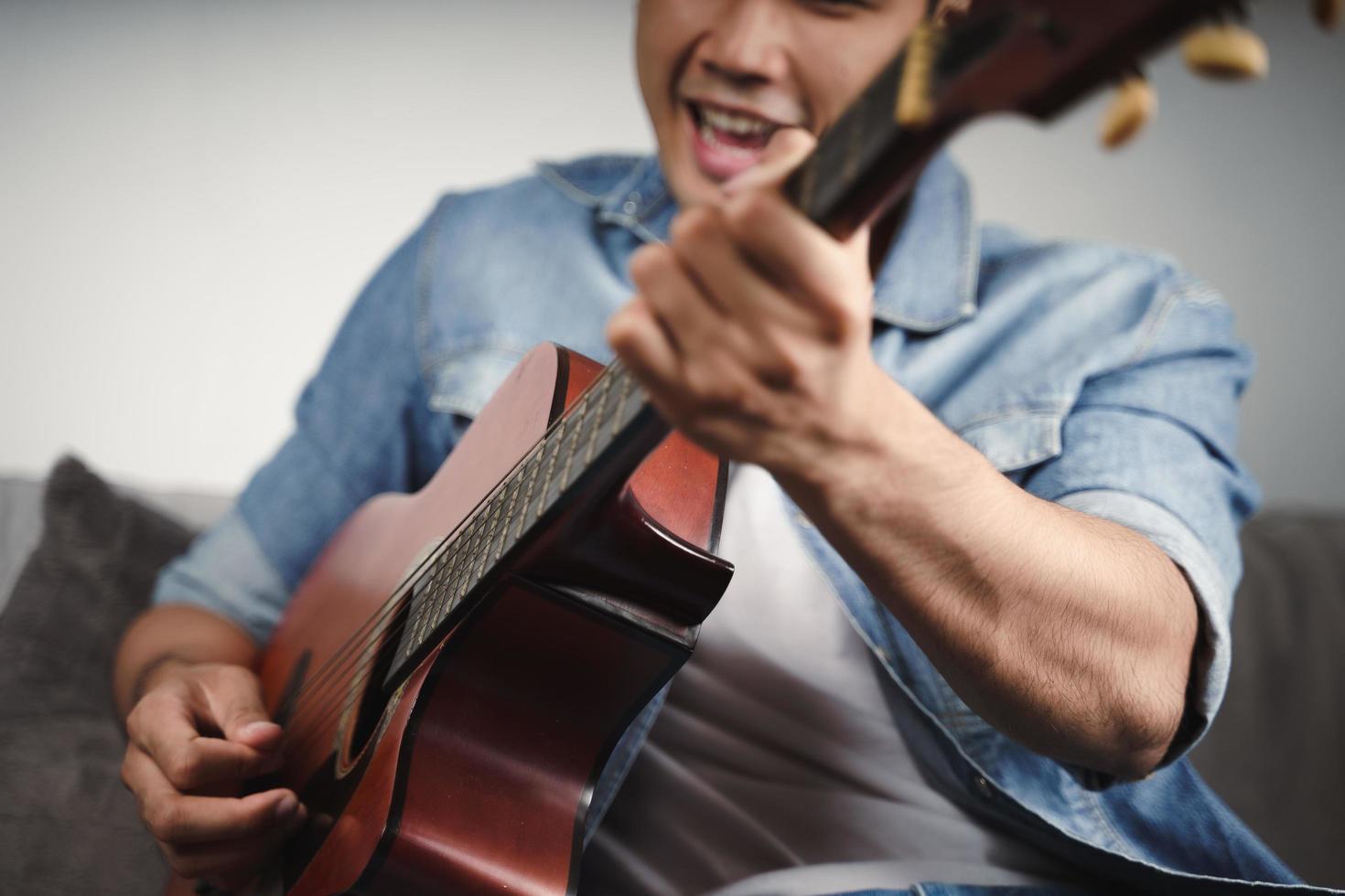 goditi un bell'uomo asiatico che si esercita o suona la chitarra sul divano del soggiorno foto