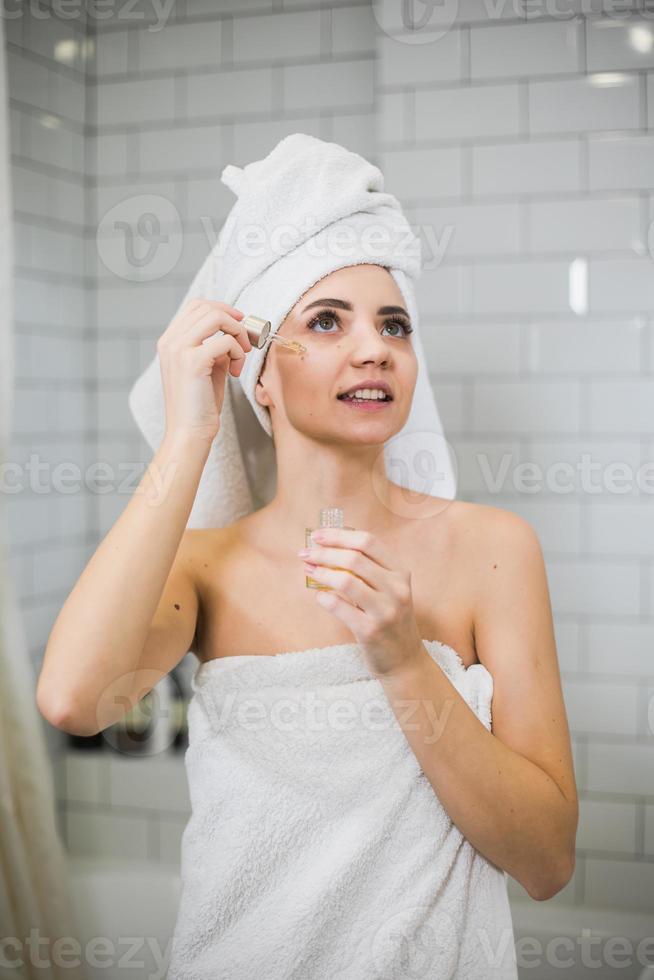 giovane donna in un asciugamano bianco applica olio idratante sulla pelle del viso. foto