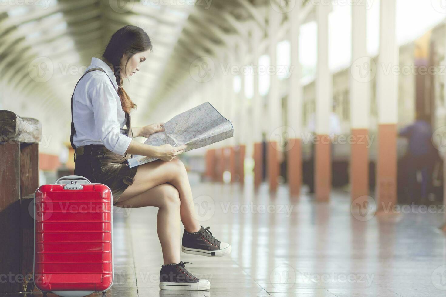 asiatico donna incinta sedersi su il panchina Guarda a il carta geografica con rosso valigia a ferrovia stazione viaggi, vacanze viaggio concetto. foto