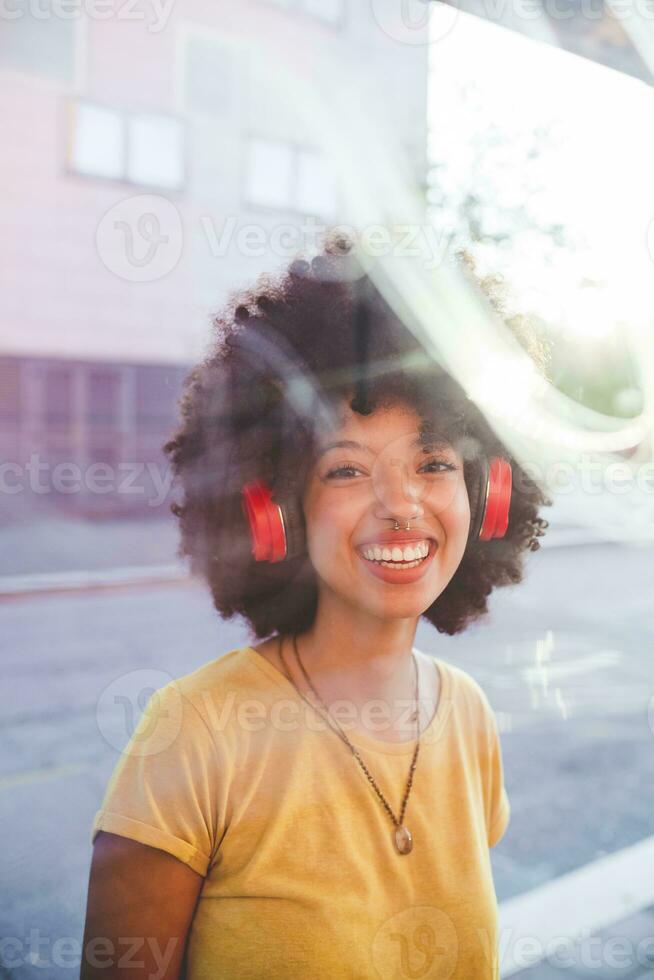 ritratto di contento giovane donna con afro pettinatura ascoltando per musica con cuffie nel il città foto
