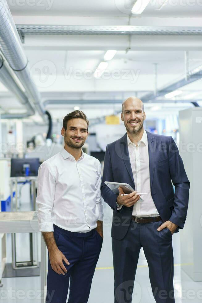sorridente maturo uomo d'affari Tenere digitale tavoletta mentre in piedi di giovane maschio ingegnere a fabbrica foto