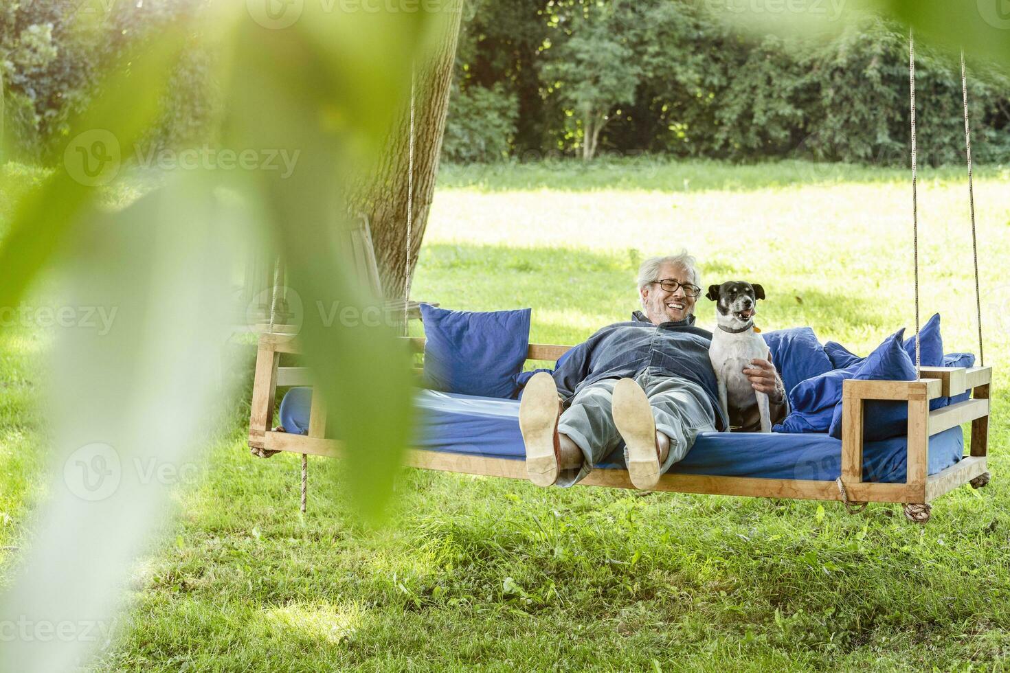 anziano uomo rilassante su un' swing letto nel il suo giardino con il suo cane foto