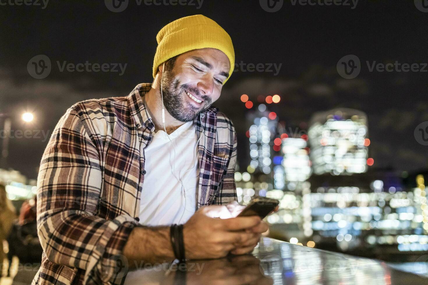 UK, Londra, sorridente uomo pendente su un' ringhiera e guardare a il suo Telefono con città luci nel sfondo foto