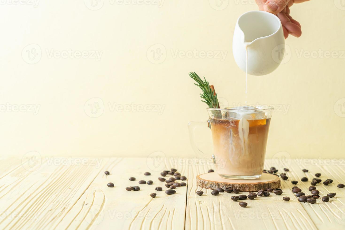 versando il latte nel bicchiere di caffè nero con cubetto di ghiaccio, cannella e rosmarino su fondo di legno foto