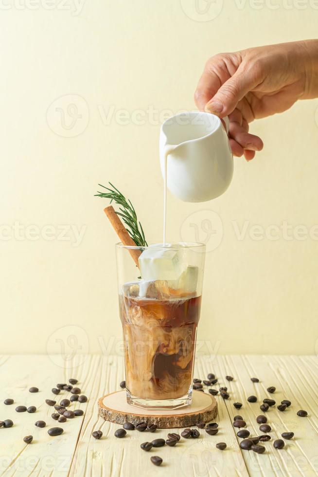 versando il latte nel bicchiere di caffè nero con cubetto di ghiaccio, cannella e rosmarino su fondo di legno foto