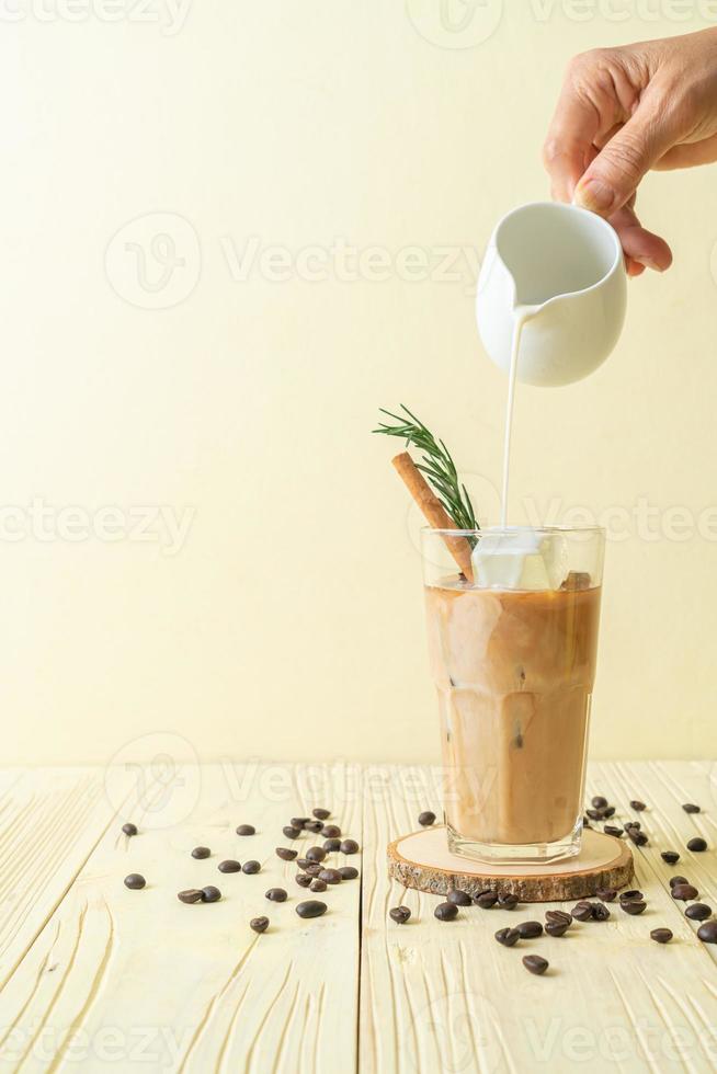versando il latte nel bicchiere di caffè nero con cubetto di ghiaccio, cannella e rosmarino su fondo di legno foto